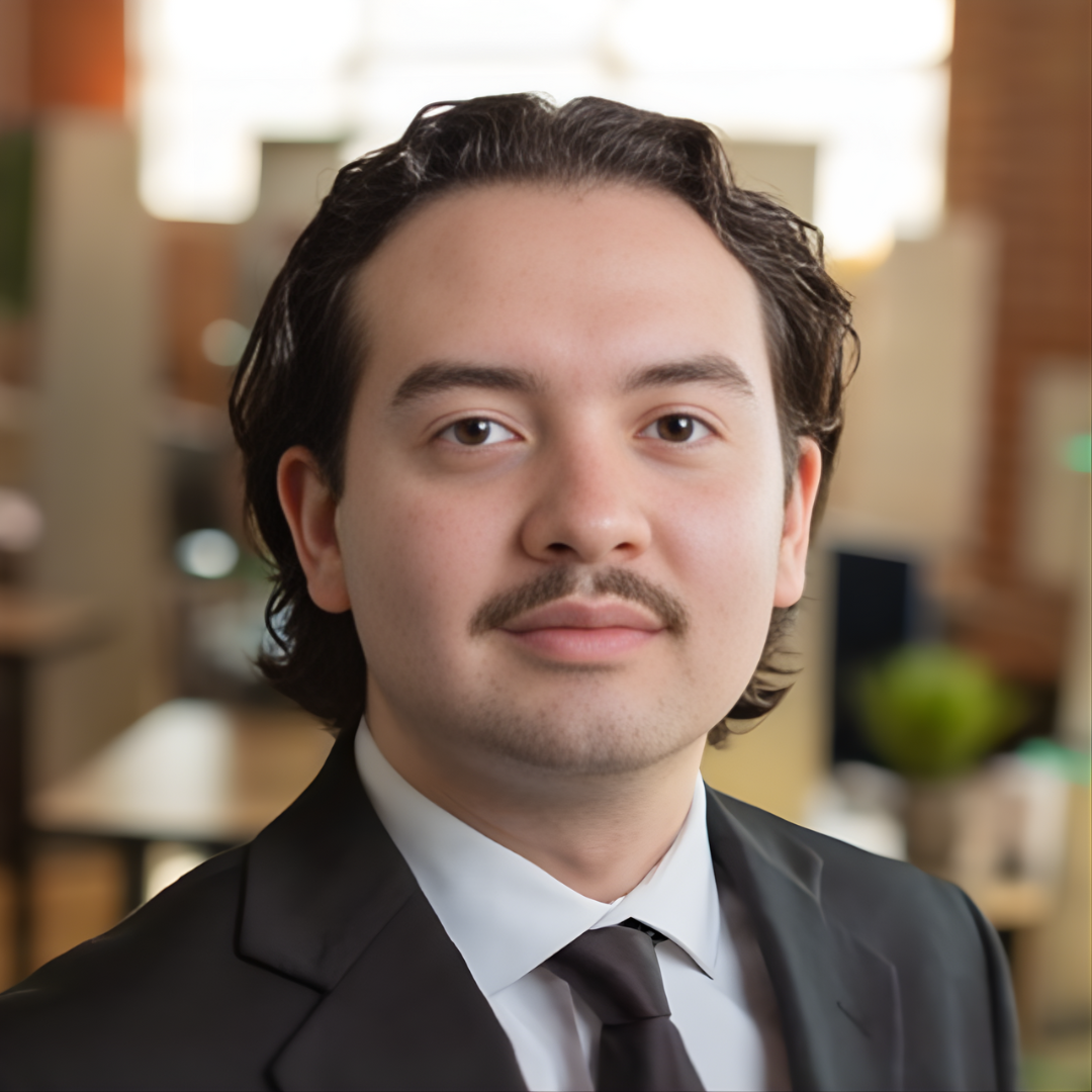 Portrait of a young man with dark wavy hair, light facial hair, wearing a black suit, white shirt, and black tie, in an office environment with blurred background.