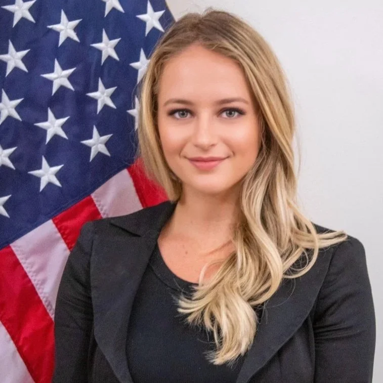 Young woman with long blonde hair smiling, standing in front of an American flag.