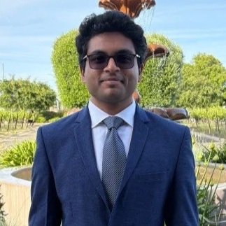 Man in a blue suit and sunglasses standing outdoors in a garden