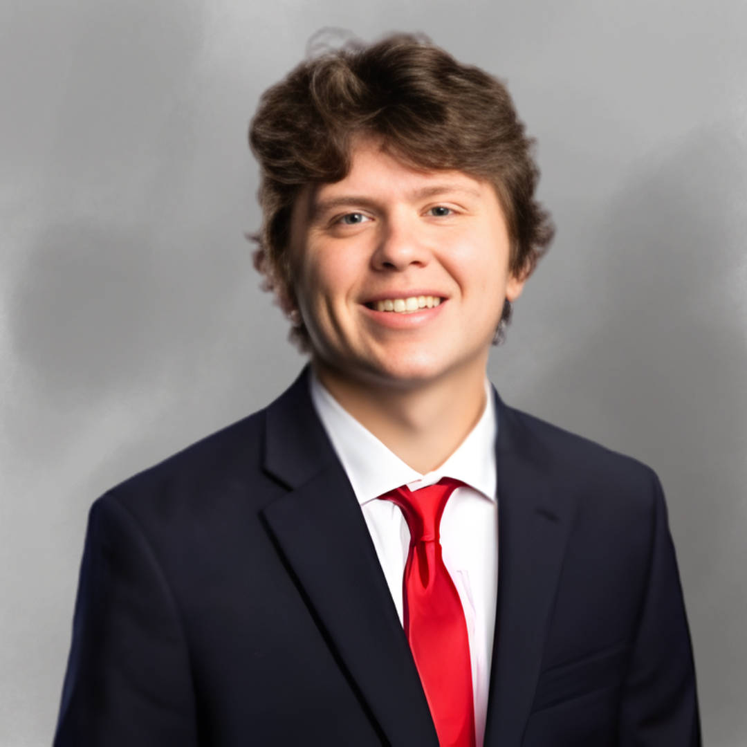 Young man in a dark suit, white shirt, and red tie smiling in front of a neutral gray background.