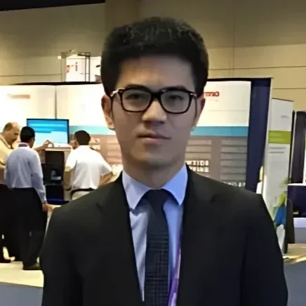 Young man in a suit and glasses standing at a conference or expo, with booths and other people in the background.