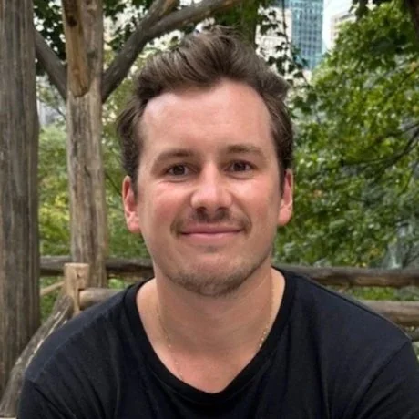 A man with short brown hair and a light beard, smiling in an outdoor park with greenery and trees.