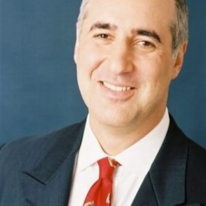A professional portrait of a smiling man in a suit and red tie, standing against a blue background.