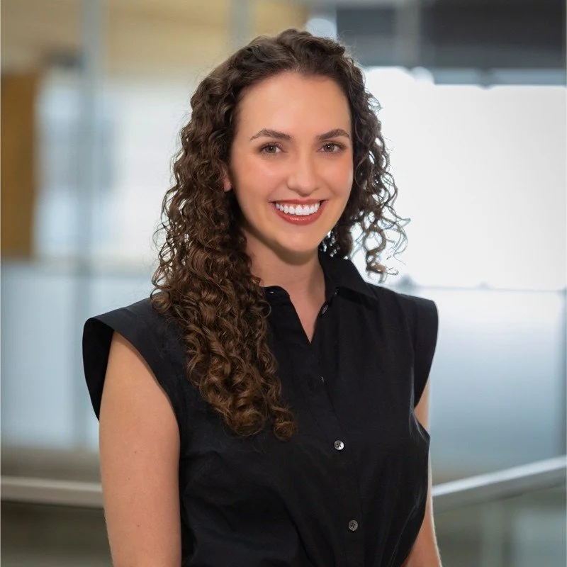 A young woman with long curly brown hair, smiling, wearing a black sleeveless blouse, standing in an office environment.