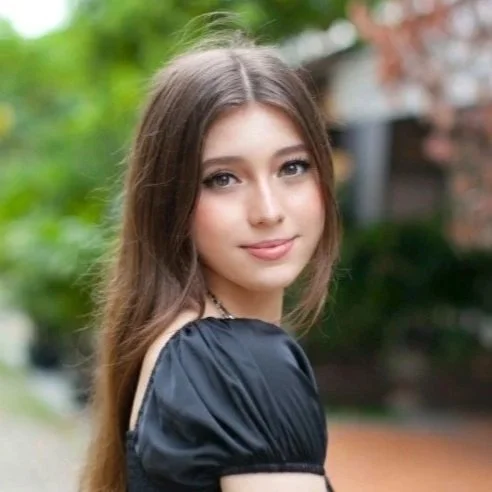 A young woman with long brown hair, wearing a black top, standing outdoors with blurred green trees and houses in the background.