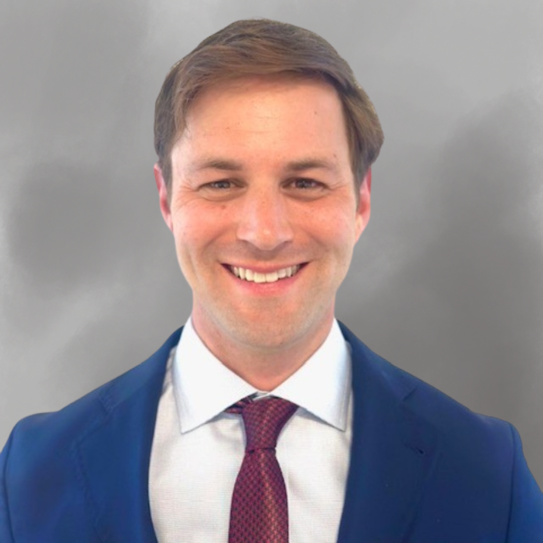 A smiling man in a navy suit, white shirt, and maroon tie, standing against a blurred gray background.