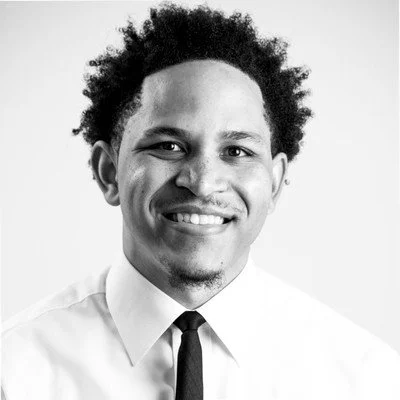 Black man with curly hair smiling, wearing a white shirt and dark tie, against a plain background.