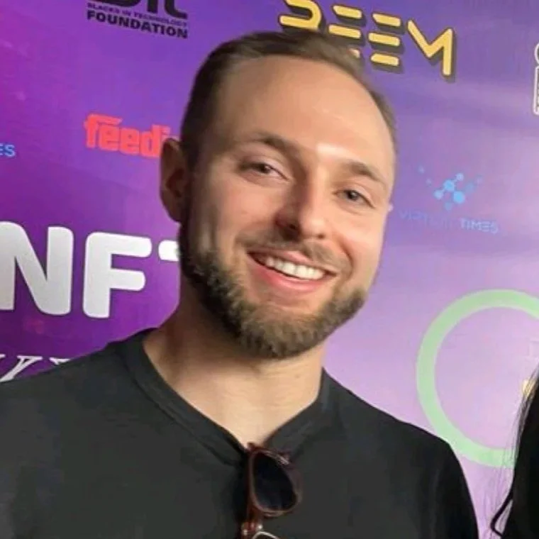 A smiling man with short hair and a beard, wearing a black shirt and sunglasses hanging from his collar, standing in front of a purple background with various logos.