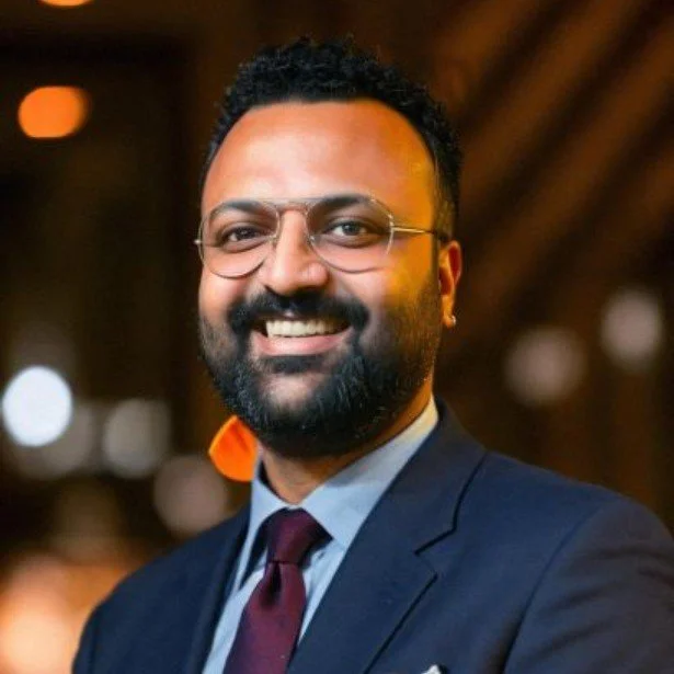 A smiling man with a beard and glasses wearing a dark suit, light blue shirt, and maroon tie, in a warmly lit indoor setting.
