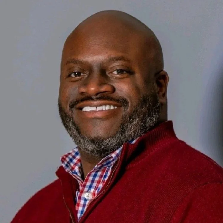 A smiling Black man with a beard, dressed in a checked shirt and a maroon sweater, posing against a gray background.