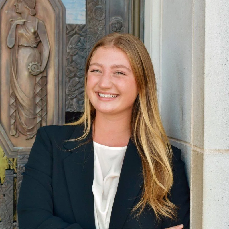 A young woman with long blonde hair, wearing a white blouse and a black blazer, standing outside near a decorative metal sculpture and wall.