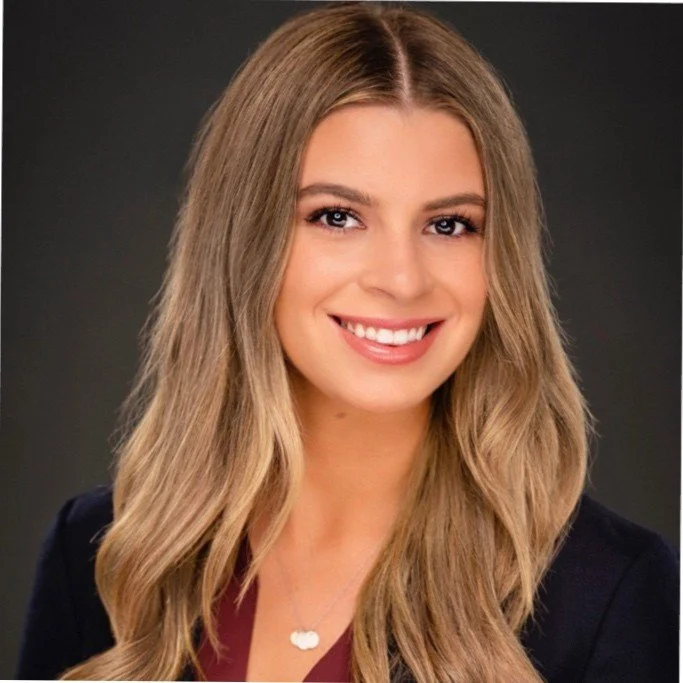 A young woman with long, wavy blonde hair smiling at the camera, wearing a dark blazer and a necklace with a cloud-shaped pendant against a dark gray background.