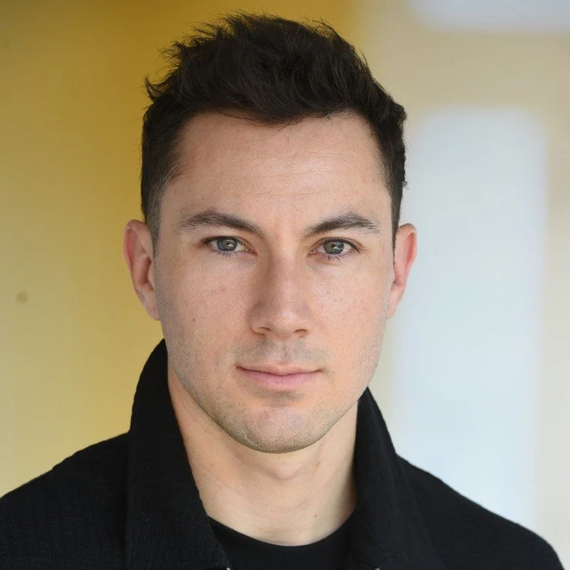 Close-up portrait of a young man with short dark hair, light skin, and subtle makeup, wearing a black shirt, looking directly at the camera with a neutral expression.