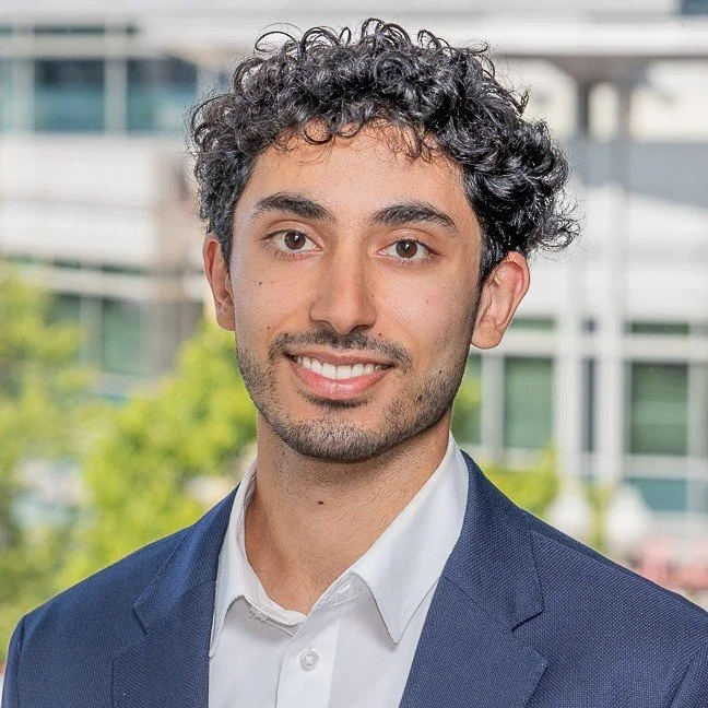 A young man with curly dark hair, wearing a white shirt and blue blazer, smiling in an outdoor setting with modern buildings in the background.