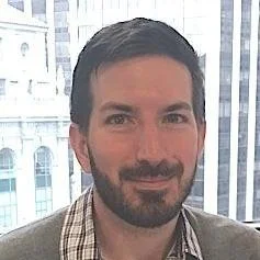 Portrait of a man with dark hair and a beard, smiling, with a cityscape view of tall buildings in the background.