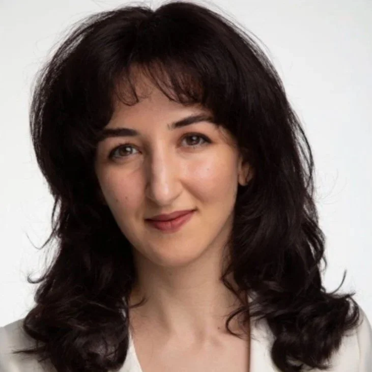 A woman with shoulder-length dark brown hair and light skin, smiling slightly, wearing a beige blazer, against a white background.
