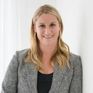portrait of a woman with blonde hair, wearing a gray checked blazer and a black top, smiling against a white background.