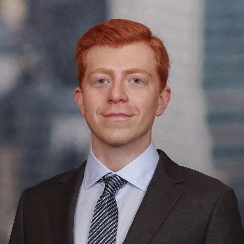 A young man with red hair wearing a black suit, white shirt, and striped tie, smiling in front of a blurred cityscape background.