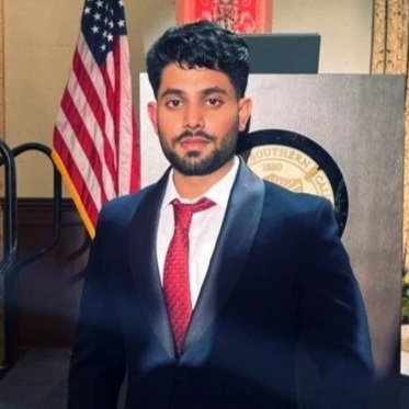 A young man in a dark suit and red tie standing in front of an American flag and a university emblem.