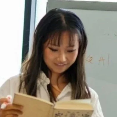 A young woman with long dark hair holding an open book, standing in front of a whiteboard.