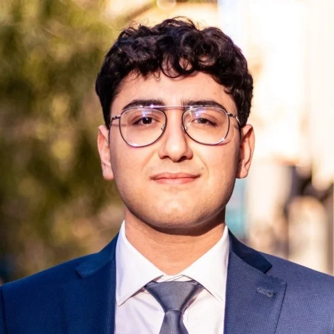 A young man with curly dark hair and glasses wearing a navy blue suit, white shirt, and gray tie outdoors with blurred green and tan background.