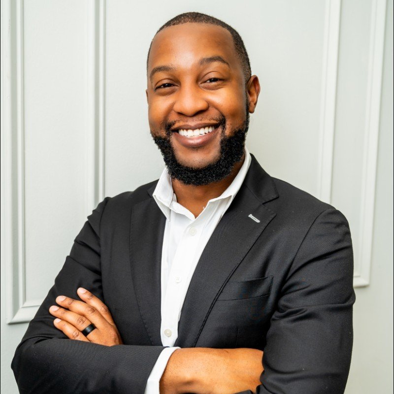 A smiling Black man with a beard, wearing a white shirt and black blazer, standing in front of a white paneled wall.