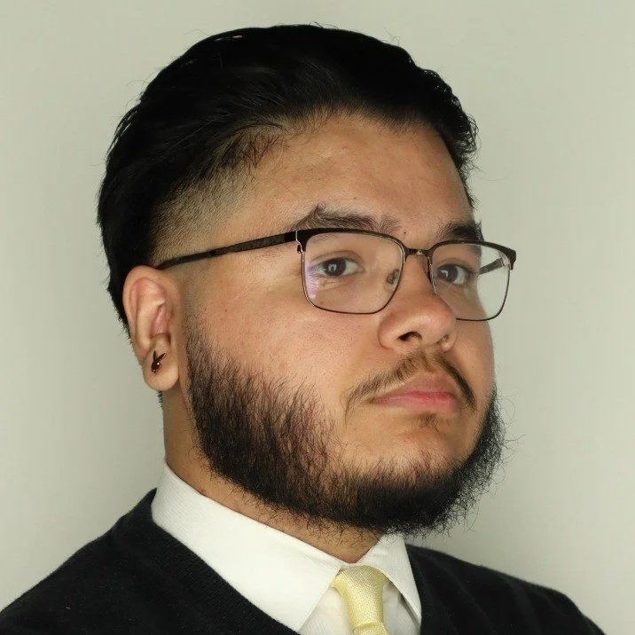 Portrait of a young man with dark hair styled back, beard, glasses, wearing a white shirt, black sweater, and light-colored tie, facing slightly to the right against a plain light background.