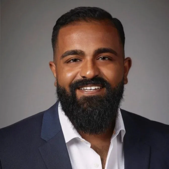 Professional headshot of a man with dark hair and beard, wearing a white shirt and dark blazer, smiling against a gray background.