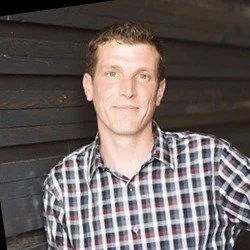 Portrait of a man with short brown hair wearing a plaid shirt, standing against a dark wooden wall.