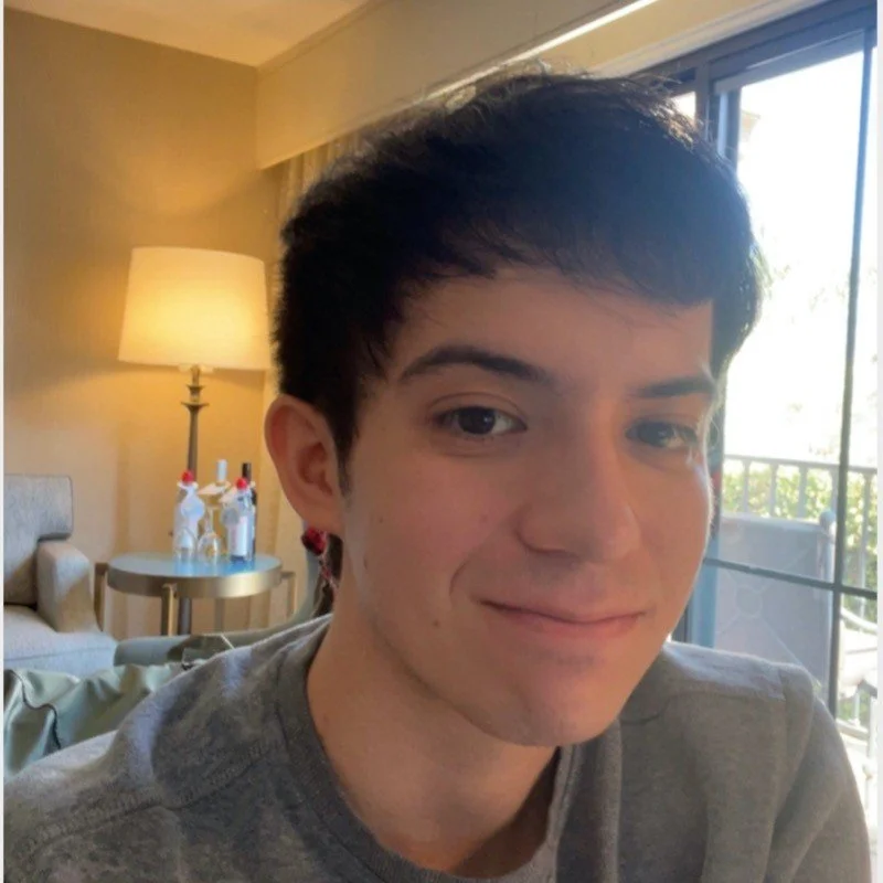 Young man with dark hair smiling, sitting in a hotel room near a sliding glass door with a view outside, beside a lamp and a table with decorative bottles.