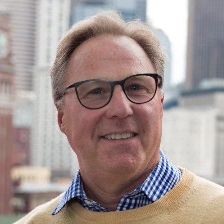 Smiling middle-aged man with glasses, wearing a blue checkered shirt and beige sweater, with city skyscrapers in the background.