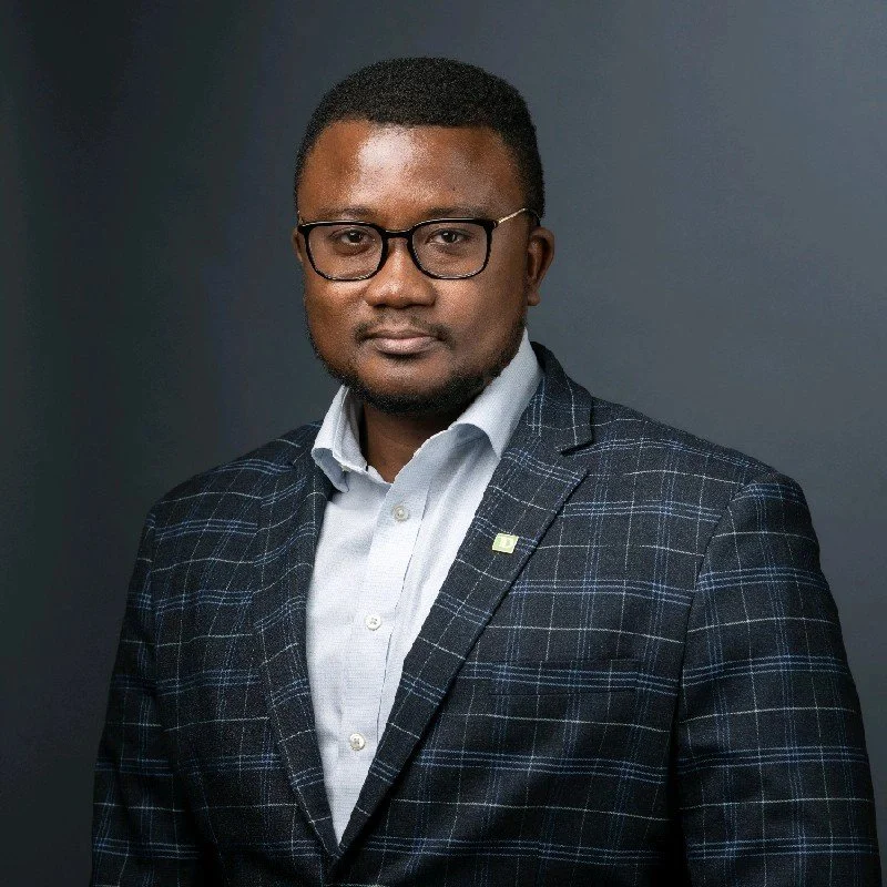 A professional headshot of a man wearing glasses, a light blue collared shirt, and a dark plaid suit jacket, standing against a dark background.