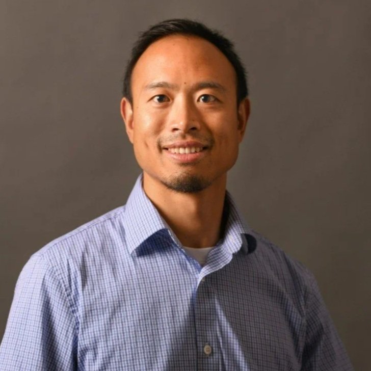 Portrait of an Asian man with short black hair, smiling, wearing a light blue checkered button-up shirt, standing against a plain gray background.