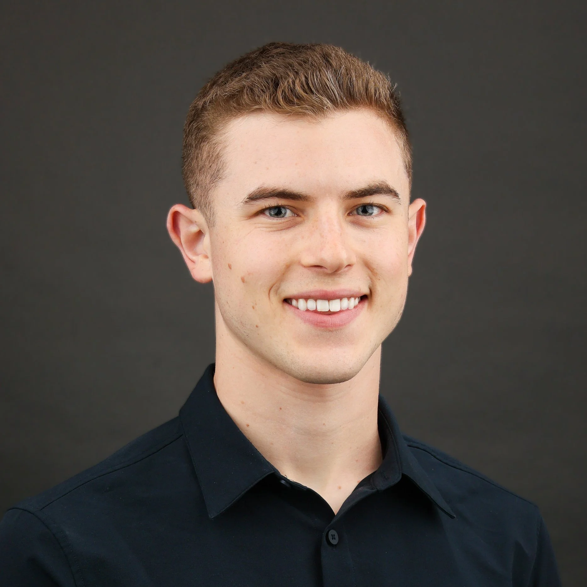 Headshot of a young man with short red hair, blue eyes, light skin, and freckles, smiling, wearing a black collared shirt against a dark gray background.