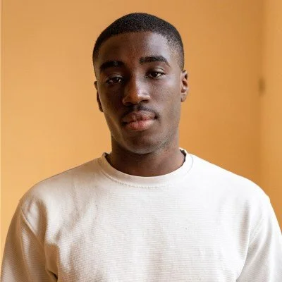 A young man with short black hair and a slight smile, wearing a white sweatshirt, standing in front of a plain, warm-colored wall.