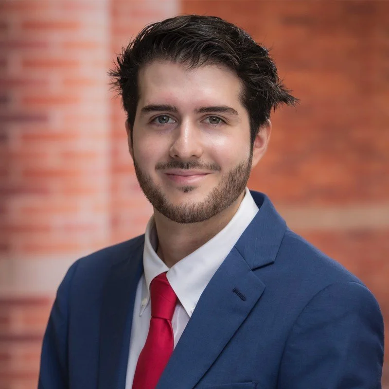 A young man in a blue suit, white shirt, and red tie, smiling with a blurred brick wall background.