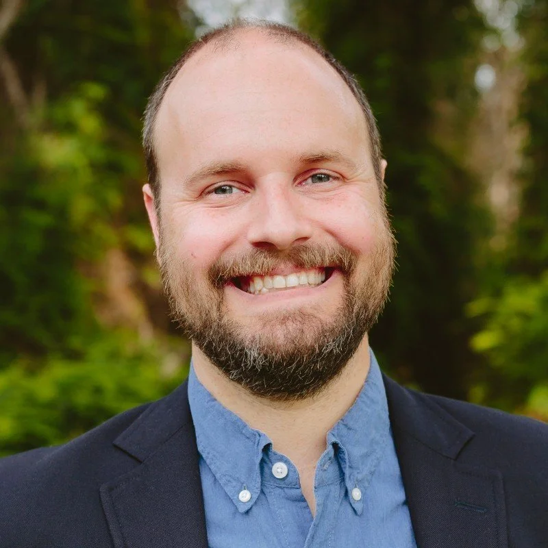 A smiling man with a beard and short hair, wearing a blue collared shirt and a dark blazer, outdoors with a blurred green forest background.