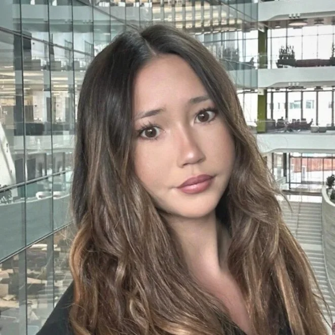 A young woman with long, wavy brown hair and natural makeup posing in a modern glass building interior.