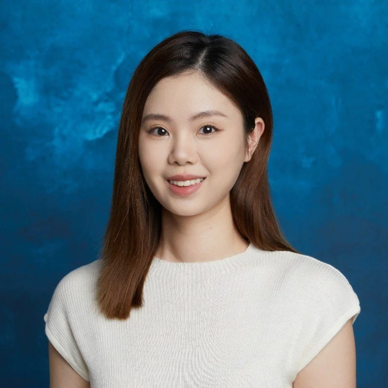 Portrait of a young Asian woman with shoulder-length brown hair, smiling, wearing a white top, against a blue textured background.