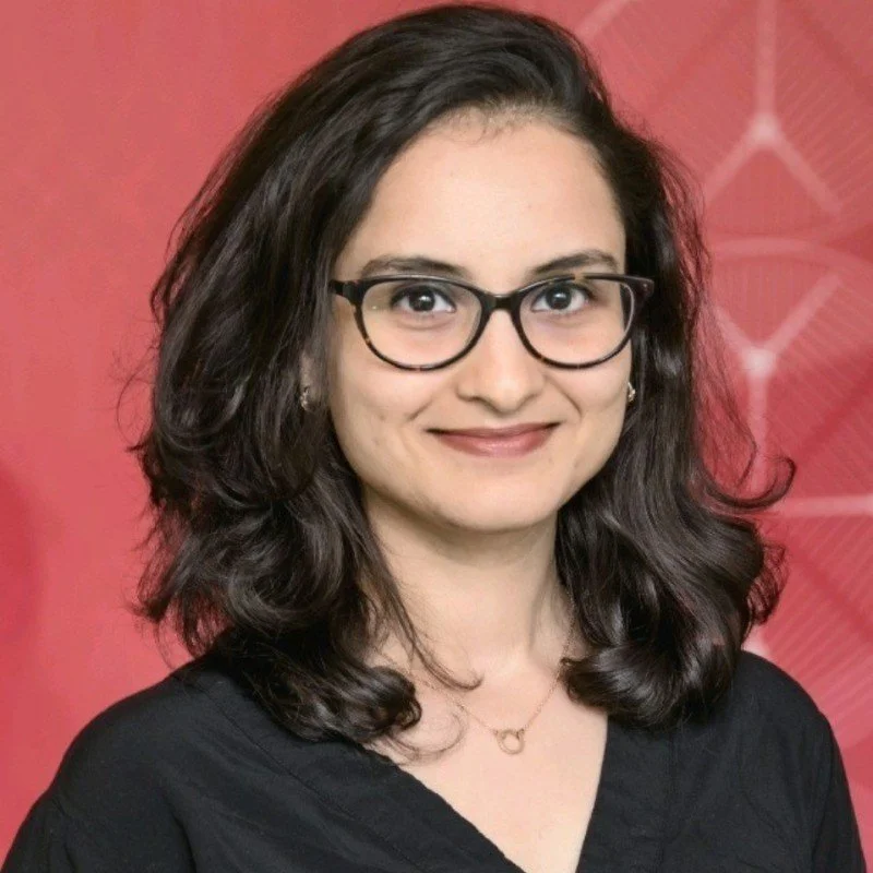 Portrait of a woman with shoulder-length dark hair, glasses, and a black top, smiling against a red geometric-patterned background.