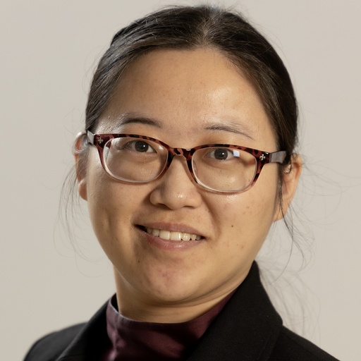 Close-up portrait of an Asian woman wearing glasses, a black blazer, and a maroon top, smiling at the camera.