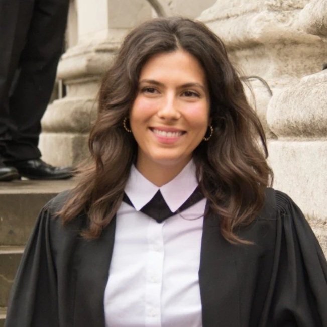 A young woman in a black graduation gown and white shirt, smiling outdoors on steps near stone columns.
