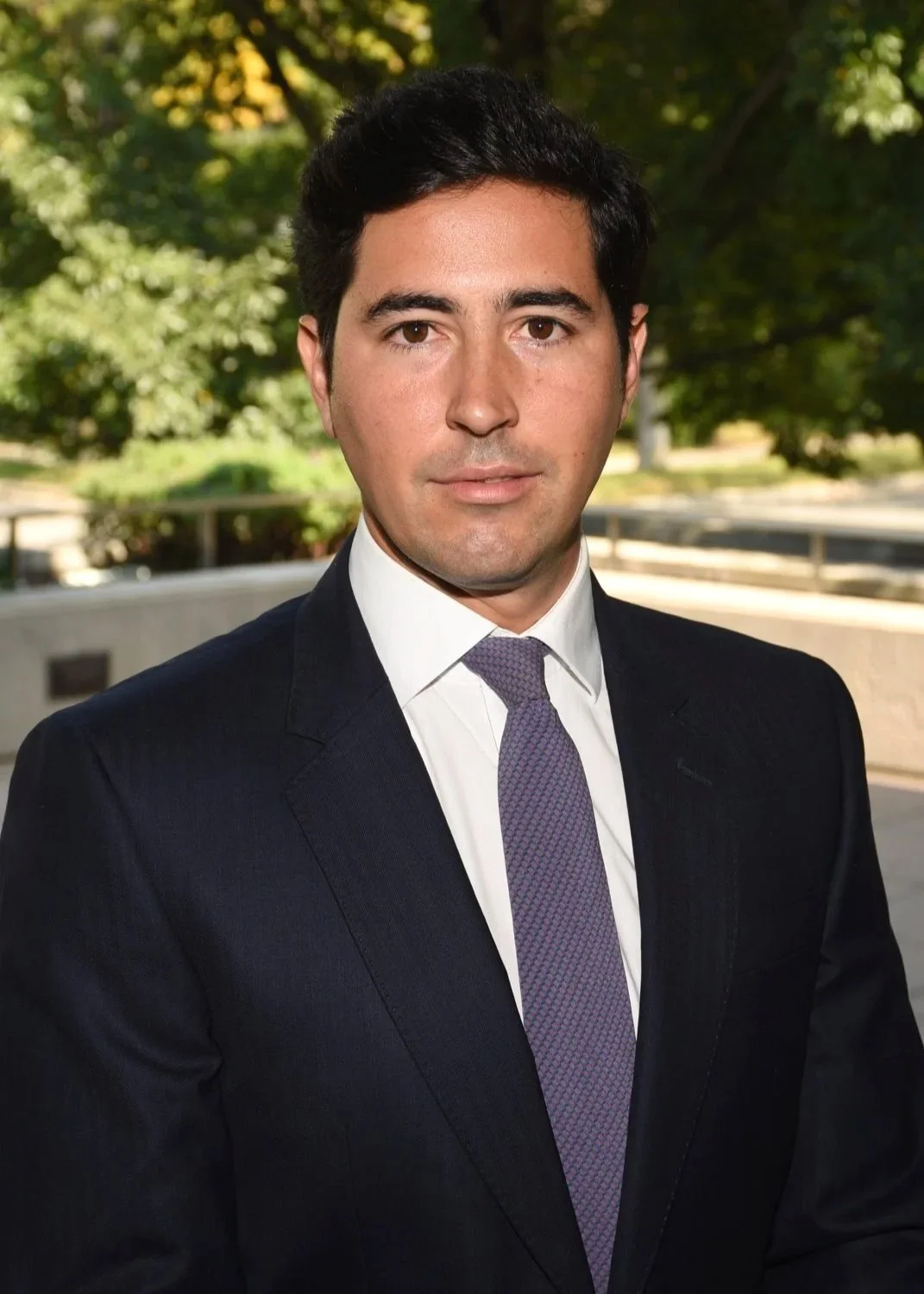 A man in a dark suit and tie standing outdoors with trees and a walkway in the background.