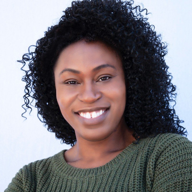 A smiling woman with curly black hair wearing a green sweater against a light background.