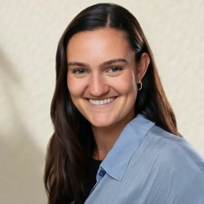 A young woman with long brown hair, smiling, wearing a light blue collared shirt and hoop earrings.
