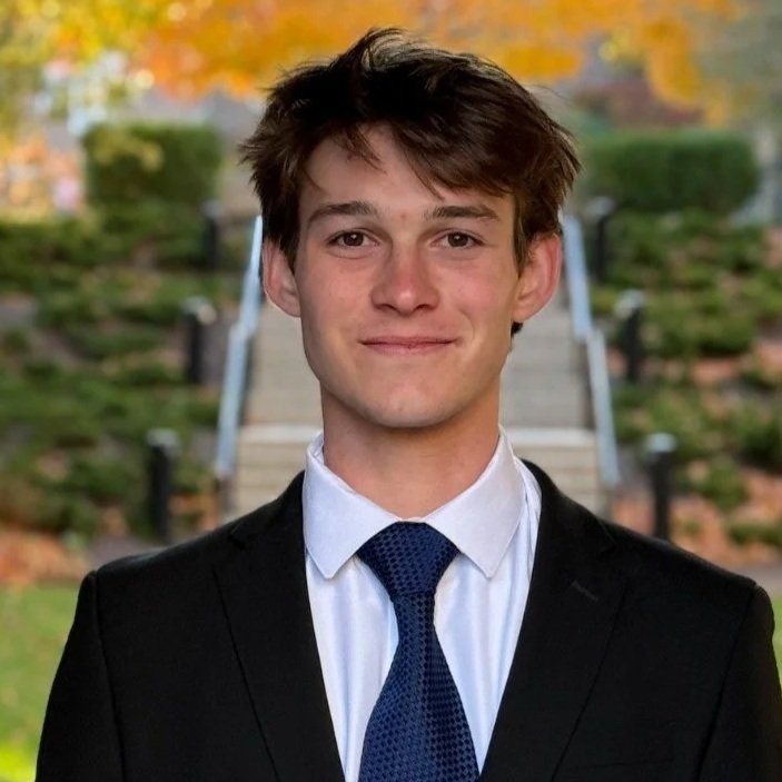Young man with brown hair smiling, wearing a black suit, white shirt, and navy blue tie, outdoors with autumn trees and steps in the background.