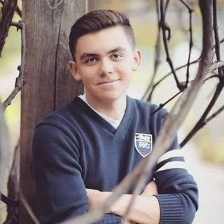 Young man with short brown hair wearing a navy blue sweater with a white shirt underneath, standing outdoors next to a tree.