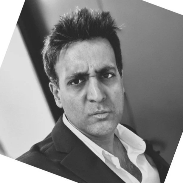 A black and white close-up portrait of a man with short, tousled hair, wearing a suit jacket and collared shirt, making a serious facial expression.