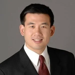 Professional headshot of a man in a black suit, white shirt, and red tie, smiling against a gray background.