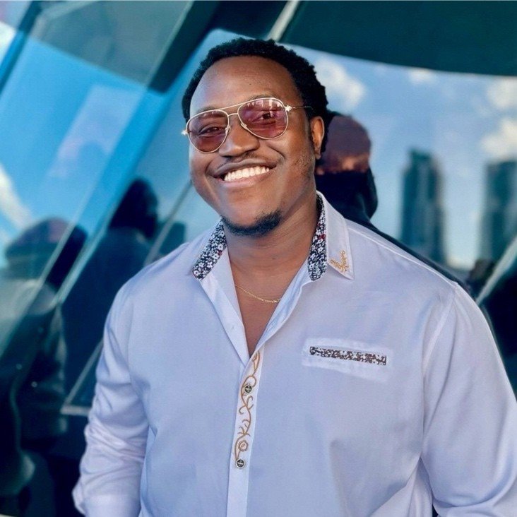 A smiling man wearing sunglasses, a light-colored shirt with decorative details on the collar and pocket, and a gold necklace, standing outdoors in front of a reflective glass building.
